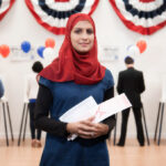 A young woman wearing a hijab at a voting location, holding a ballot.