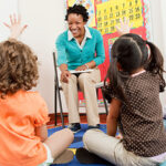 Teacher and children sitting on floors with hands raised