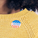 A young African American woman wears a bright yellow sweater with a Vote sticker on it