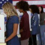 Four women of various demographics filling in ballots and casting votes in booths at polling station.
