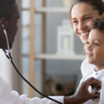A doctor holds a stethoscope to exam a child during a health care visit.